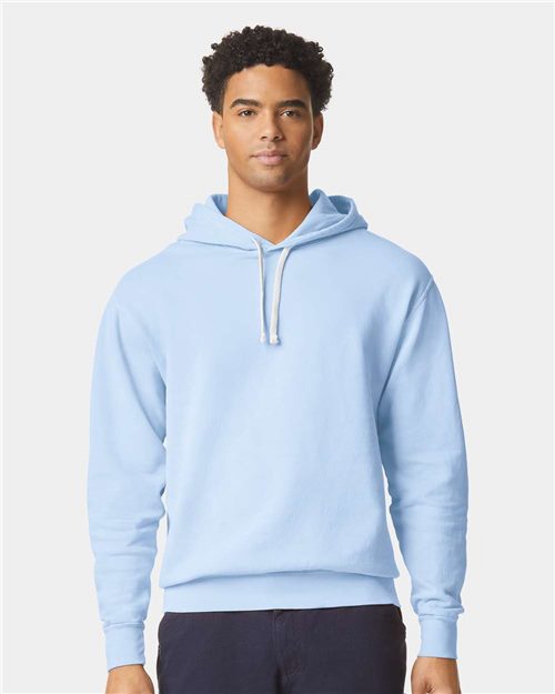 A person with short, curly hair stands against a white background wearing a Comfort Colors Garment-Dyed Lightweight Fleece Hooded Sweatshirt in light blue with a white drawstring. Made from OEKO-TEX certified fabric, it features a relaxed fit and long sleeves. They're also wearing dark pants.