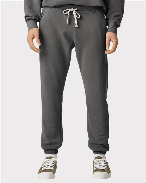 A person wears Comfort Colors - Garment-Dyed Lightweight Fleece Sweatpants in dark gray, featuring a drawstring waistband, crafted from ring-spun cotton, paired with a black sweater and white/olive green sneakers against a plain light gray background.