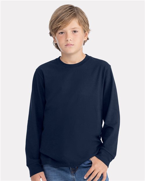 A young boy with short, light brown hair is wearing a plain dark blue Next Level - Youth Cotton Long Sleeve T-Shirt and classic fit blue jeans. He stands against a white background, looking directly at the camera with a neutral expression.