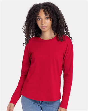 A person with curly hair wears a red Next Level Women's Cotton Relaxed Long Sleeve T-Shirt and relaxed-fit blue jeans, standing against a plain white background.