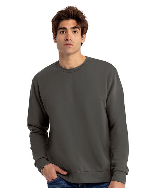 A brown-haired person wears a Next Level Santa Barbara Sweatshirt in dark gray with blue jeans, standing against a white background and looking at the camera neutrally.