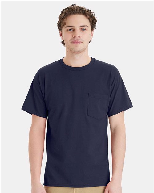 A person with short, wavy brown hair is wearing a plain navy blue Hanes Essential-T Pocket T-Shirt, crafted through socially conscious manufacturing. They are standing against a light gray background.