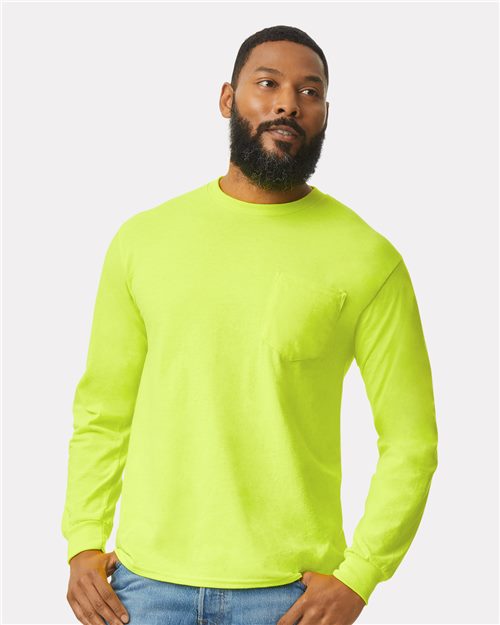 A man with a beard wears a bright yellow Gildan Ultra Cotton® Long Sleeve Pocket T-Shirt, made from 100% cotton, paired with blue jeans. The shirt's chest pocket pops against the plain light background.