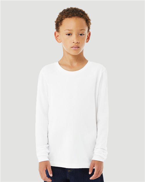 A young boy wearing a BELLA + CANVAS - Youth Heather CVC Long Sleeve Tee, made from airlume combed cotton, stands against a plain gray background. His hands rest at his sides, and he has short curly hair.