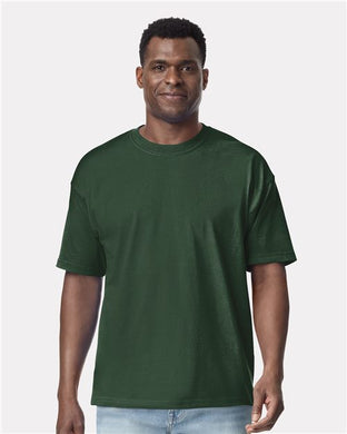 A person in a plain dark green Gildan - Hammer™ Maxweight T-Shirt and light blue jeans stands against a white background, smiling slightly while looking forward, highlighting the quality of ring-spun cotton.