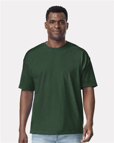A person in a plain dark green Gildan - Hammer™ Maxweight T-Shirt and light blue jeans stands against a white background, smiling slightly while looking forward, highlighting the quality of ring-spun cotton.