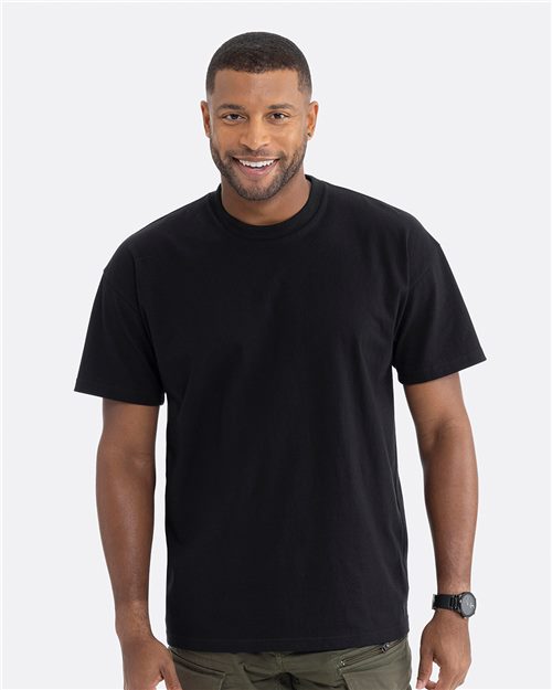 A man smiles confidently, wearing a Next Level - Heavyweight T-Shirt in plain black, made of combed ring-spun cotton, paired with olive green pants against a light gray background. The outfit's dropped shoulders and relaxed fit are completed with a stylish black wristwatch.