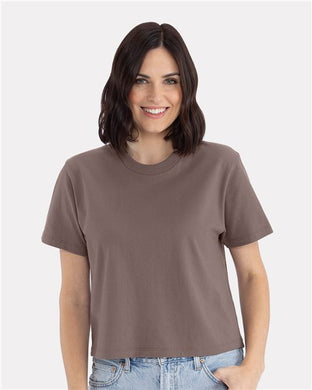 A woman with shoulder-length dark hair smiles in a Next Level Women's Heavyweight Boxy T-Shirt, wearing a short-sleeve brown tee in a relaxed fit made of combed ring-spun cotton and light blue jeans. The background is light gray.