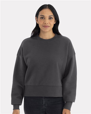 A person with long dark hair wears the relaxed fit, dark gray Next Level Women's Heavyweight Crewneck Sweatshirt made from recycled polyester. They stand facing forward against a plain light gray background.