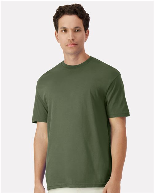 A man with short, dark hair wears a Gildan Light Cotton T-Shirt in olive green and light pants. Standing against a white backdrop, he looks ahead with a neutral expression.