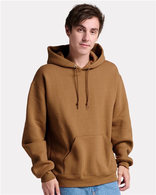 A person stands against a white background, hands in the front pocket of their brown JERZEES - Rugged Hooded Sweatshirt. Made from sustainably-sourced cotton, this cozy JERZEES pullover features heavyweight fleece for lasting comfort.