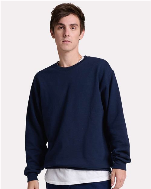 A person with short brown hair wears a JERZEES Ultimate CVC Crewneck Sweatshirt in navy blue, made from sustainably-sourced cotton, over a white shirt. They stand against a plain light gray background, facing the camera with a neutral expression.