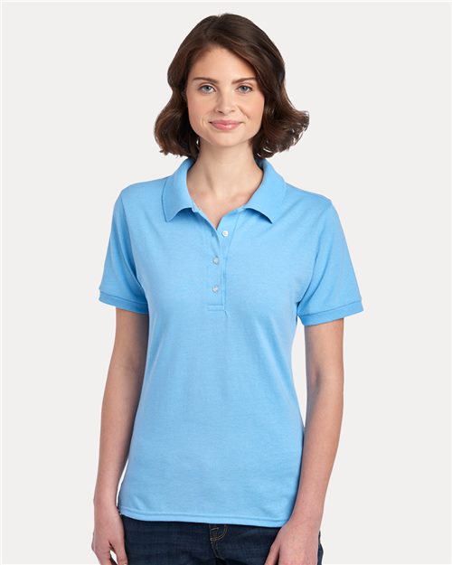 A person with shoulder-length brown hair wears a JERZEES Women's Dri-Power® Polo in light blue against a plain white background, smiling slightly with relaxed arms, enjoying the comfort and moisture-management features of the cotton/polyester blend shirt.