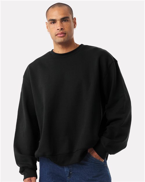 Load image into Gallery viewer, Against a plain white background, a person with a shaved head confidently stands in the BELLA + CANVAS - 10 oz. Heavyweight Sweatshirt crafted from Airlume combed cotton, paired with blue jeans, one hand casually in their pocket, showcasing style blended with sustainable manufacturing.
