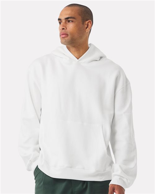 Load image into Gallery viewer, A man in a plain white BELLA + CANVAS - 10 oz. Heavyweight Hoodie and green pants glances sideways against a simple backdrop, highlighting the unisex silhouette made with Airlume combed cotton for exceptional softness and crafted sustainably.
