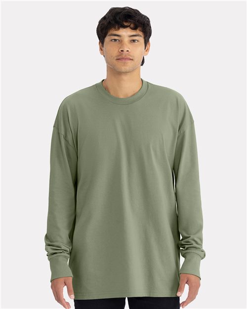 Someone wearing a Next Level Heavyweight Long Sleeve T-Shirt in olive green, made of combed ring-spun cotton, stands against a light gray background, looking directly at the camera with a neutral expression.