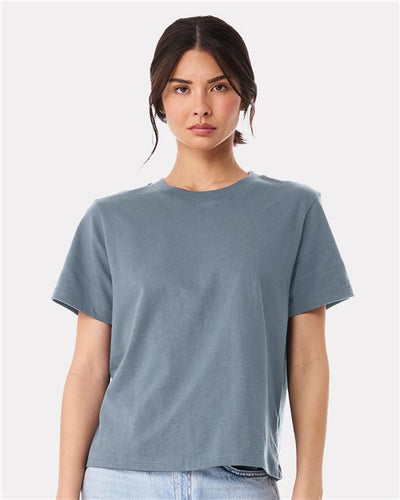 A person with long dark hair wears a plain BELLA + CANVAS Women's 6 oz Heavyweight Tee and light blue jeans, standing against a white background and looking straight at the camera, reflecting sustainable manufacturing.