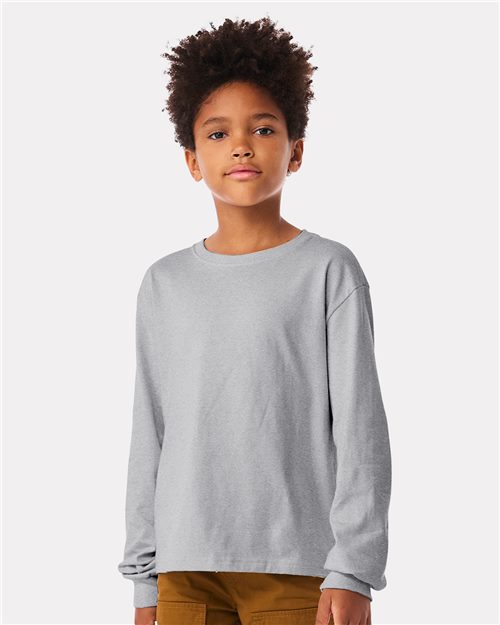 Cheerful child in a BELLA + CANVAS Youth 6oz. Heavyweight Long Sleeve Tee and brown pants, standing against a simple, light-colored backdrop.