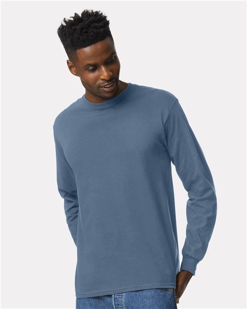 A man in a Gildan Ultra Cotton® long-sleeve T-shirt, blue and made from sustainable cotton, paired with denim jeans, glances downward with a slight smile against a plain light gray background.