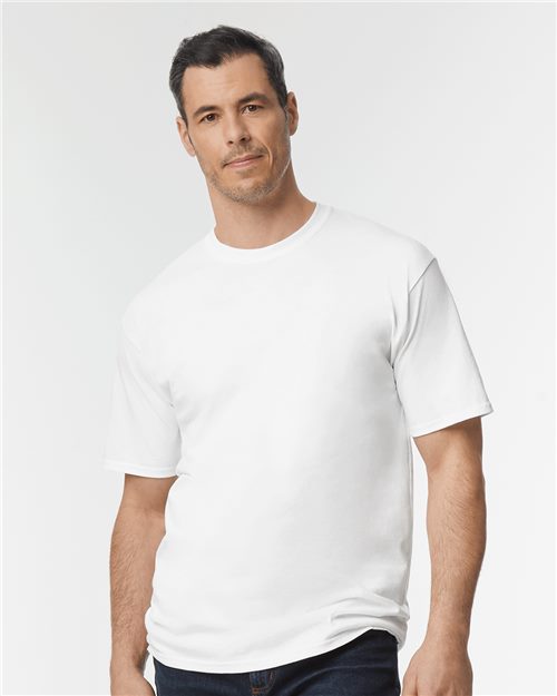 A man with short dark hair and a beard wears a Gildan Ultra Cotton® Tall T-Shirt in white and dark jeans against a light gray background, his neutral expression and the relaxed positioning of one hand by his side highlighting his support for sustainable manufacturing.