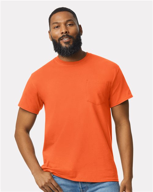 A bearded person smiles with their hands at their sides, wearing blue jeans and an orange Gildan Ultra Cotton® Pocket T-Shirt made from 100% cotton. The backdrop is plain light gray.