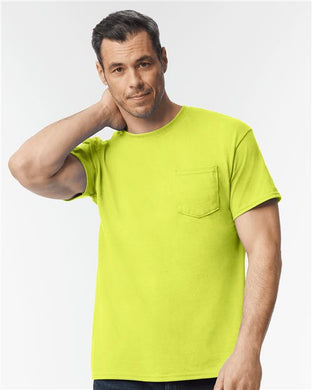 A man with short dark hair stands against a white background, wearing a bright yellow-green Gildan DryBlend® Pocket T-Shirt made from a cotton/polyester blend. His hand on the back of his neck emphasizes the garment's commitment to sustainable manufacturing.