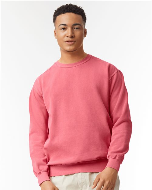 A person wearing a Comfort Colors Garment-Dyed Sweatshirt in pink and beige pants stands against a plain white backdrop. With short, curly hair and a neutral expression, they embody socially conscious manufacturing through sustainable attire.