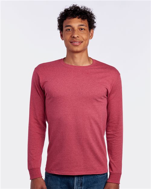 A person with curly hair is smiling while wearing a JERZEES - Dri-Power® Long Sleeve 50/50 T-Shirt and blue jeans, standing against a plain white background.