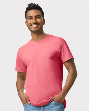 A person stands against a plain light gray background, smiling in a Gildan - Heavy Cotton™ T-Shirt in bright pink and blue jeans, with hands in pockets.