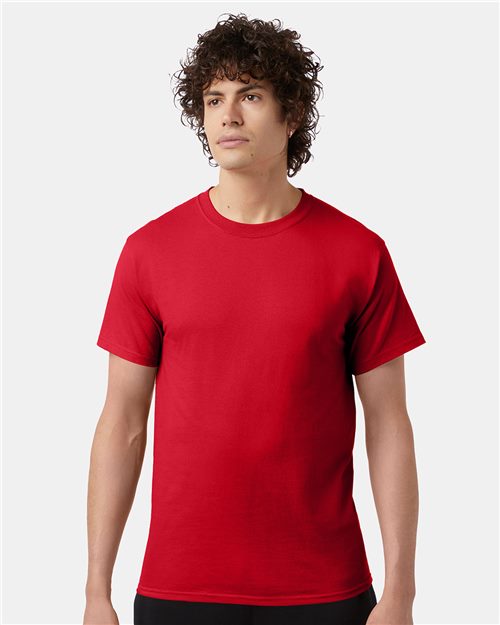 A person with curly hair wears a Champion T-Shirt from Champion, in plain red cotton, standing against a plain gray background and looking slightly to the side.