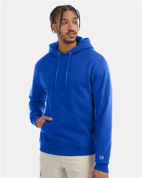 A person in a bright blue Champion Powerblend® Hooded Sweatshirt made from recycled polyester, with a front pocket and drawstrings, stands against a plain gray background. They have short braided hair and are looking to the side, wearing beige pants.