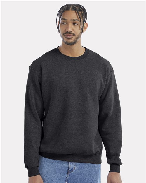 Load image into Gallery viewer, Wearing a dark gray Champion Powerblend® Crewneck Sweatshirt with blue jeans, the person stands against a light gray backdrop. Their neutral expression and braided hair embody Sustainable Style as they gaze slightly to the side.
