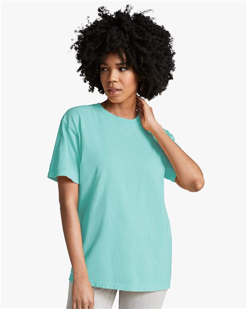 Load image into Gallery viewer, A woman with curly hair, wearing a turquoise Comfort Colors Garment-Dyed Heavyweight T-shirt made from ring-spun cotton and light-colored pants, stands against a plain white background, looking to the side.
