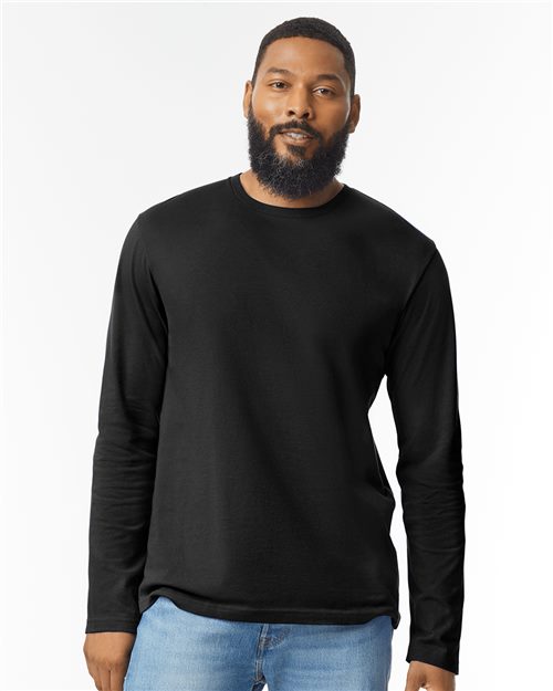 A bearded person wearing a Gildan Softstyle® Long Sleeve T-Shirt and blue jeans stands against a plain white background.