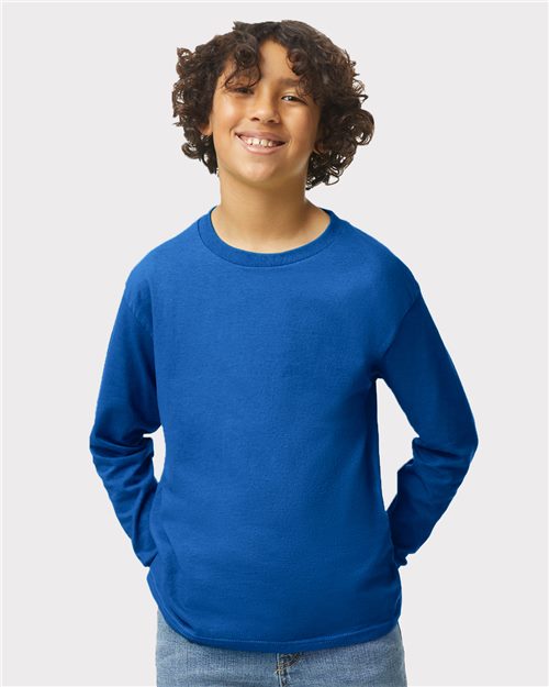 Load image into Gallery viewer, A smiling child with curly hair, wearing a bright blue Gildan Heavy Cotton™ Youth Long Sleeve T-Shirt and jeans, stands against a plain light background with hands in pockets.
