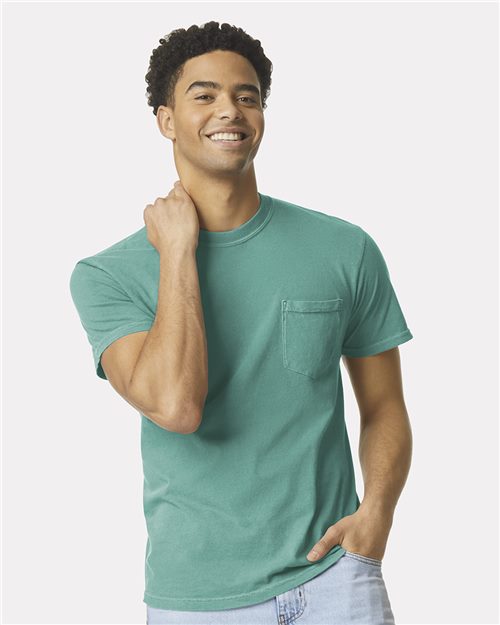 A man smiles in a Comfort Colors Garment-Dyed Heavyweight Pocket T-Shirt, made from sustainable ring-spun cotton, paired with light blue jeans. His short curly hair complements the plain white background, reflecting the brand's eco-friendly focus as he rests a hand on his neck.