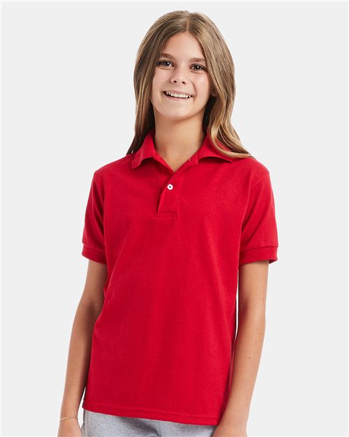 A young person with long hair wears a bright red Hanes Youth Ecosmart® Jersey Polo made from CiCLO polyester. Standing against a light gray background with relaxed arms, they embody sustainable style.