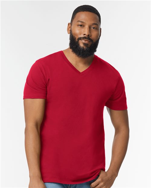 A bearded person with short hair wears a Gildan - Softstyle® V-Neck T-Shirt in bright red, made from OEKO-TEX certified ring-spun cotton, paired with blue jeans. They stand against a plain white background, looking directly at the camera with a relaxed expression.