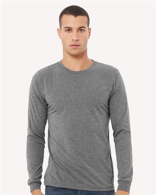 A person in a unisex BELLA + CANVAS Triblend Long Sleeve Tee, dyed with Blue Sign certified dyes, stands facing forward against a plain white background. The fitted gray shirt highlights unisex sizing while the individual has short hair and a neutral expression.