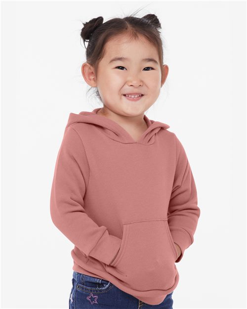 A young child with dark buns smiles, donning a pink BELLA + CANVAS - Toddler Sponge Fleece Pullover Hoodie made of Airlume combed cotton. Their hands rest in the front pocket against a plain white backdrop, emphasizing this soft and sustainably crafted garment's adherence to California EPA regulations.