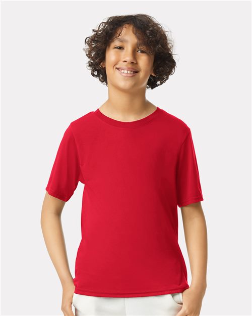 A smiling child with curly hair stands against a plain background, wearing a Gildan - Performance® Youth T-Shirt in bright red, made from sustainably crafted polyester known for its moisture-management properties, paired with light-colored pants.