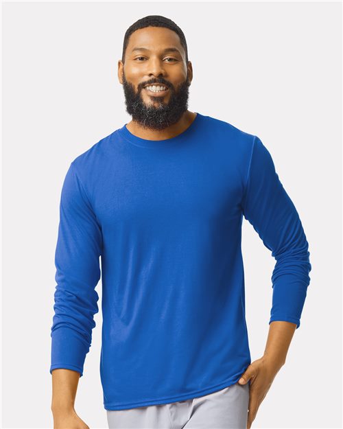 A man with a beard smiles while wearing a Gildan Performance® Long Sleeve T-Shirt in bright blue, made of moisture-management polyester, paired with light gray pants against a plain white background.