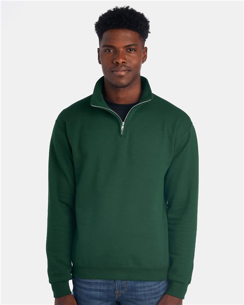 Load image into Gallery viewer, Against a plain white background, an individual with short curly hair exudes calm confidence in a dark green JERZEES Nublend® Cadet Collar Quarter-Zip Sweatshirt made of pill-resistant fleece over a black shirt. Blue jeans complete the casually stylish look.
