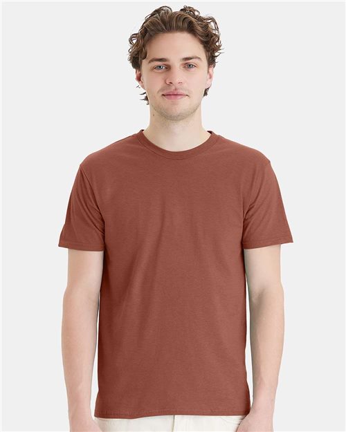 A person with curly brown hair wears a plain, short-sleeved Hanes Perfect-T T-shirt made from ring-spun cotton and light-colored pants. They stand against a gray background, looking directly at the camera with a neutral expression.