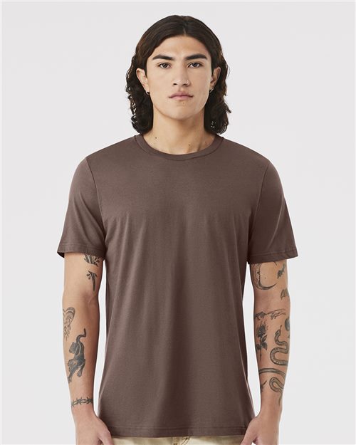 A person with long, dark hair faces forward wearing a plain brown BELLA + CANVAS - Jersey Tee made from Airlume combed cotton. Arm tattoos are visible as they stand neutrally against a light gray background. The unisex sizing of the shirt ensures it's versatile for all.