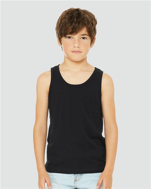 A child stands against a grey background, wearing the BELLA + CANVAS - Youth Jersey Tank made from Airlume combed cotton and light blue jeans. With short brown hair and a calm expression, they showcase unisex sizing and the sustainable fashion promise of BELLA + CANVAS.