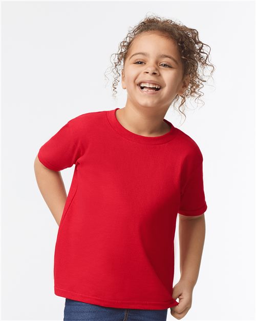 A curly-haired child joyfully smiles, wearing a bright red Gildan - Heavy Cotton™ Toddler T-Shirt made from 100% U.S. cotton paired with blue jeans against a light backdrop.