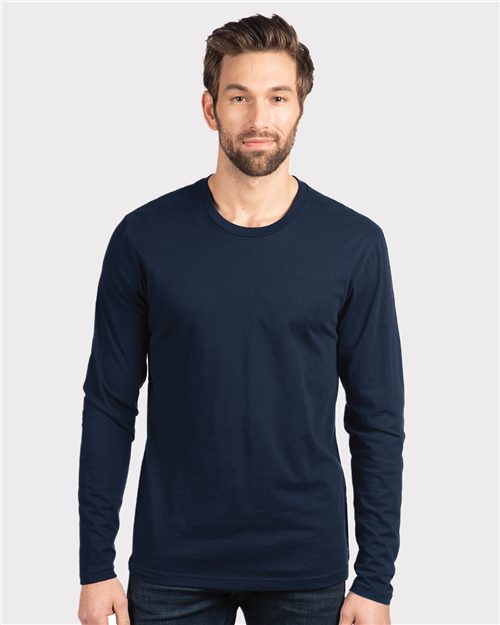 A bearded man in a plain, navy Next Level - Cotton Long Sleeve T-Shirt, crafted from combed ring-spun cotton, stands against a light gray background. His short, styled hair and neutral expression highlight an understated sustainable style.