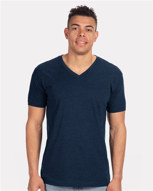 A person stands against a plain white background, wearing the Next Level CVC V-Neck T-Shirt in dark blue, crafted from fabric-laundered cotton/polyester, and light blue jeans. Socially conscious manufacturing ensures they smile confidently, arms relaxed by their sides.