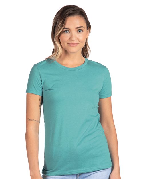 Load image into Gallery viewer, A person with shoulder-length hair in a Next Level women&#39;s CVC T-shirt and light blue jeans stands facing the camera, smiling. They have a tattoo on their right arm, representing socially conscious manufacturing. The background is plain white.
